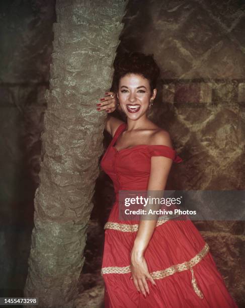 Puerto Rican singer, dancer and actress Rita Moreno, wearing a red dress with gold detail on the skirt, smiling as she leans against the trunk of a...