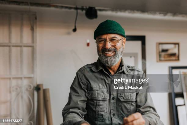 portrait of cheerful male owner wearing knit hat and eyeglasses in workshop - männer über 40 stock-fotos und bilder
