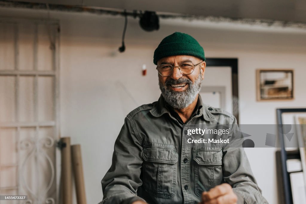 Portrait of cheerful male owner wearing knit hat and eyeglasses in workshop