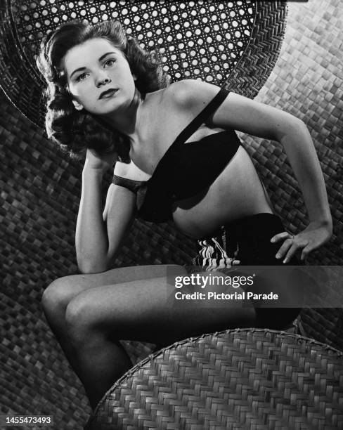 American actress Debra Paget, wearing a black two-piece swimsuit, the shoulder strap falling over her left shoulder, sitting with her right hand...