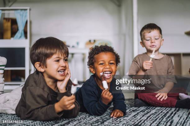 happy multiracial male students eating ice creams on carpet at day care center - gefrorene süßspeise stock-fotos und bilder