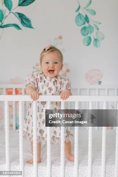 one year old girl in pink dress stands in crib and smiles - baby girls fotografías e imágenes de stock