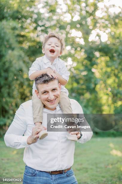 2-4 year old boy sits on top of his father's shoulders and smiles - small man and tall woman stock pictures, royalty-free photos & images
