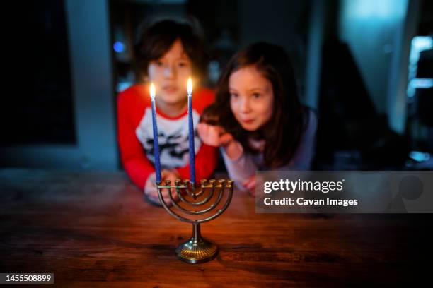 two siblings sit at table together staring at lit candles on menorah - menorah lights stock pictures, royalty-free photos & images