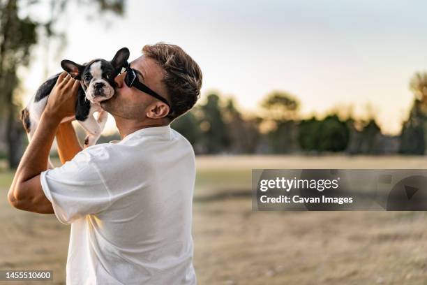 handsome young man holding up and kissing a french bulldog - puppies wearing sunglasses stock pictures, royalty-free photos & images