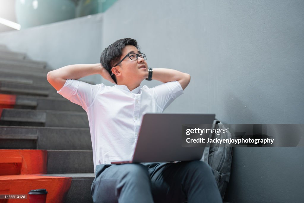 Relaxed smiling young asian businessman sitting and stretching with his laptop while taking a break