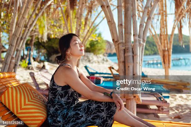 young asian woman enjoying a relaxing moment, sitting on bench by the beach - island hut stock pictures, royalty-free photos & images