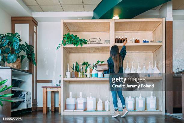 young hispanic or latina woman stocking reaching high to stock shelves with empty glass reusable bottles in zero waste store - reusable stock pictures, royalty-free photos & images