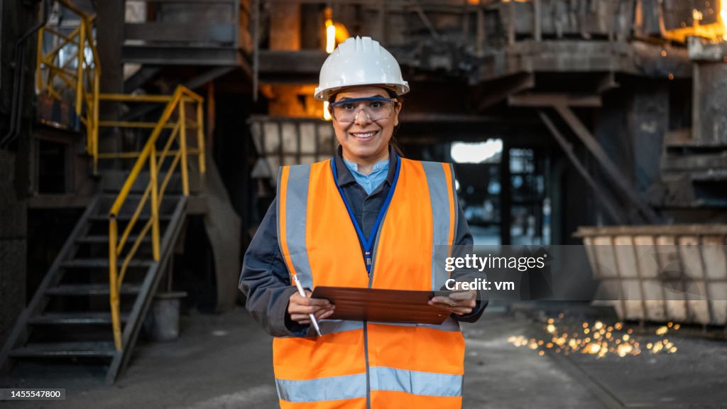 Smiling Hispanic woman in factory, female industrial worker standing in workshop, looking at camera.
