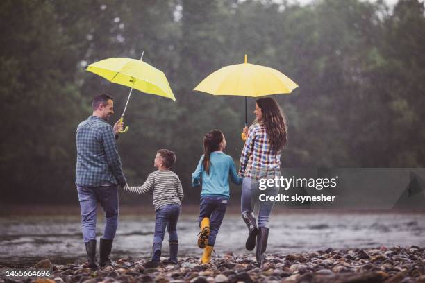 rückansicht der verspielten familie, die an einem regnerischen tag am flussufer läuft. - mother protecting from rain stock-fotos und bilder