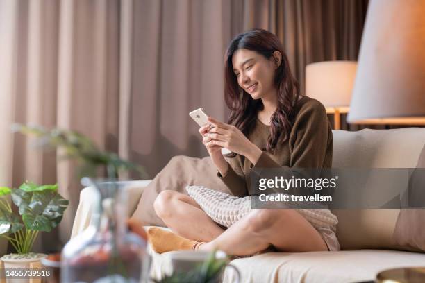 attractive young asia adult woman using a mobile phone while relaxing on the sofa at home,asian female using smartphone surfing the internet social media or chatting with her friend smiling cheerful moment at home - confident looking to camera stock pictures, royalty-free photos & images
