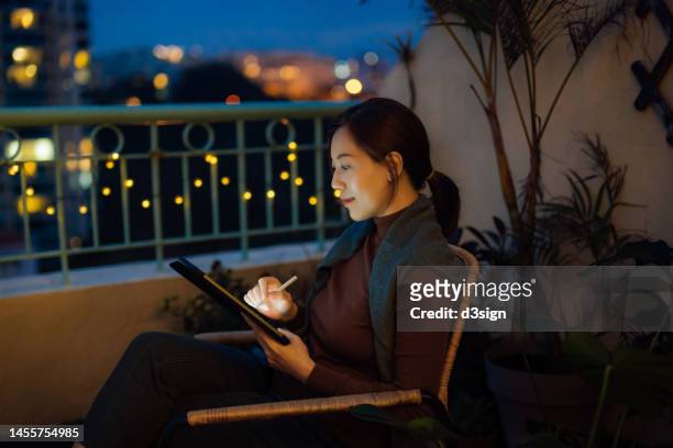 young asian woman sitting on armchair in the balcony, using digital tablet at home. surrounded by potted plants. illuminated cityscape during twilight in background. lifestyle and technology. working from home - e learning draw stock pictures, royalty-free photos & images