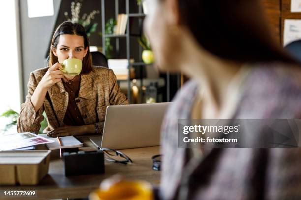 zwei geschäftsfrauen sitzen in einer reihe an ihren schreibtischen, plaudern und trinken kaffee - coffee break office stock-fotos und bilder