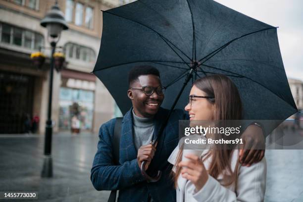 pareja joven y amorosa caminando con sombrilla y café en la ciudad - white and black women and umbrella fotografías e imágenes de stock