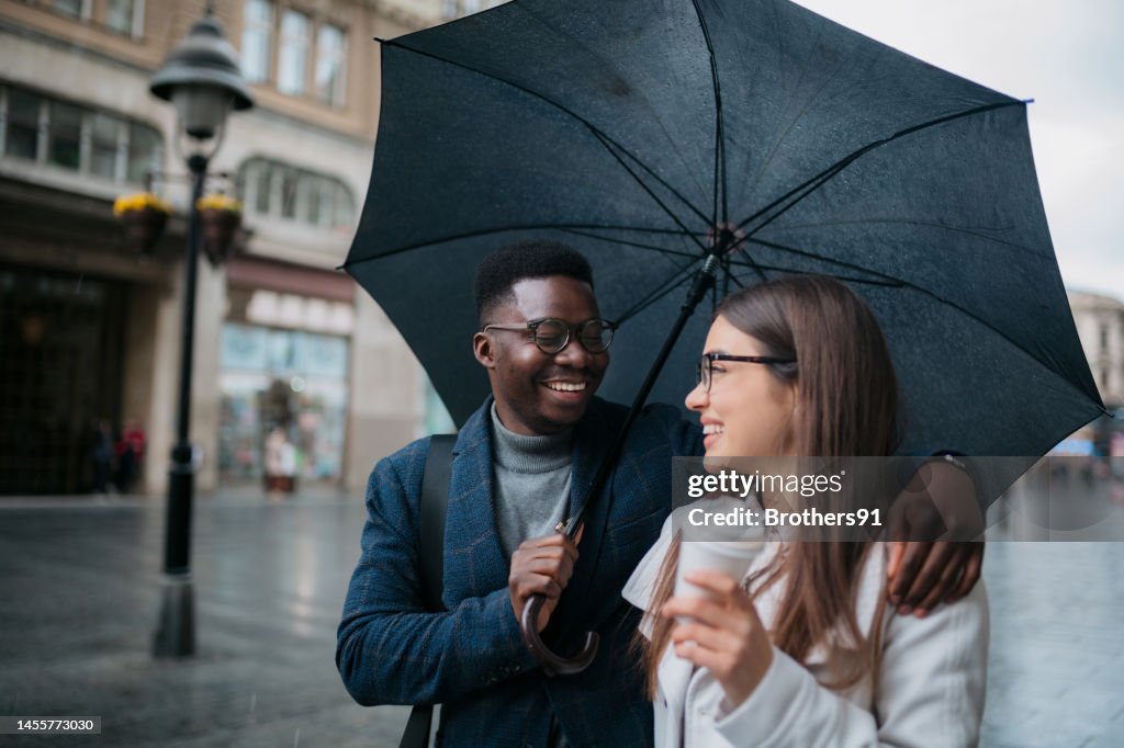 Loving young couple walking with umbrella and coffee in the city