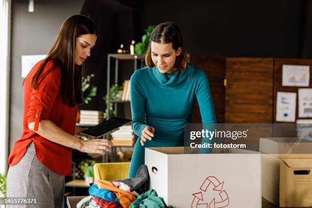 two young women folding and sorting clothes for donation and charity - vrijwilliger bedrijf stockfoto's en -beelden