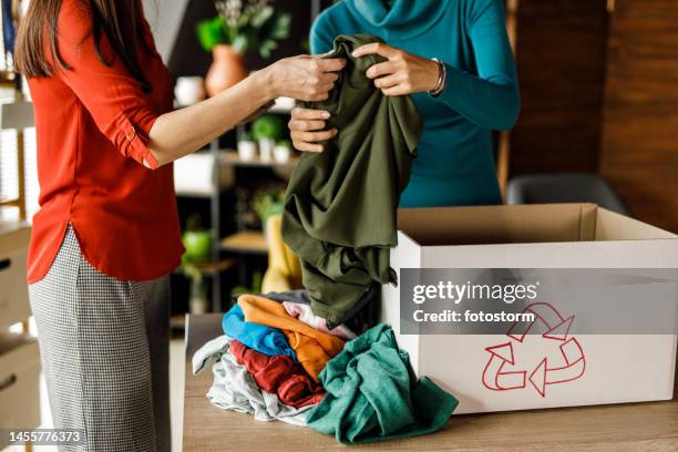 two young women folding and sorting clothes for donation and charity - textile industry stock pictures, royalty-free photos & images