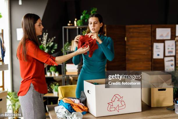 two young women folding and sorting clothes for donation and charity - clothing donation stock pictures, royalty-free photos & images