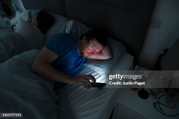 a mature man lies awake in bed whilst his partner sleeps. he uses his smartphone - smartphone lesen nacht stock-fotos und bilder