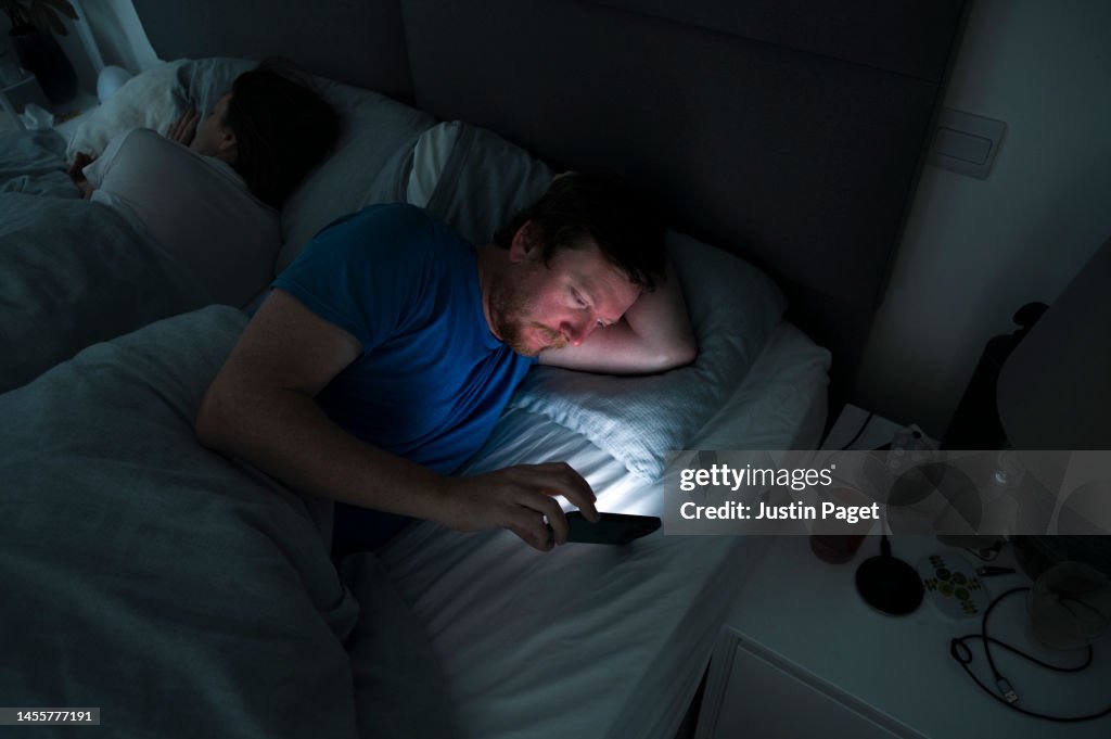 A mature man lies awake in bed whilst his partner sleeps. He uses his smartphone