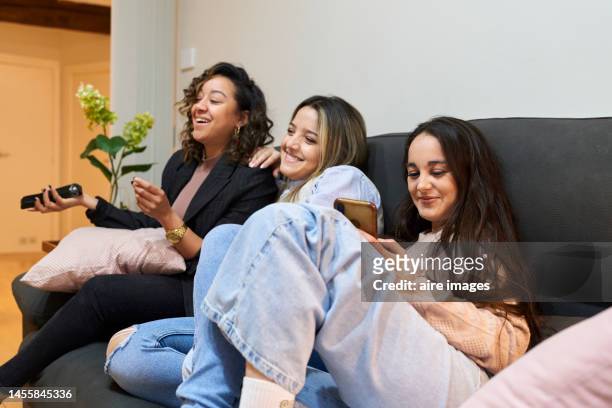 multiracial group of female friends watching tv at home, relaxing on living room sofa, laughing and chatting. friday night in city apartment, background flower vase and front door. - anticipation excited stock pictures, royalty-free photos & images
