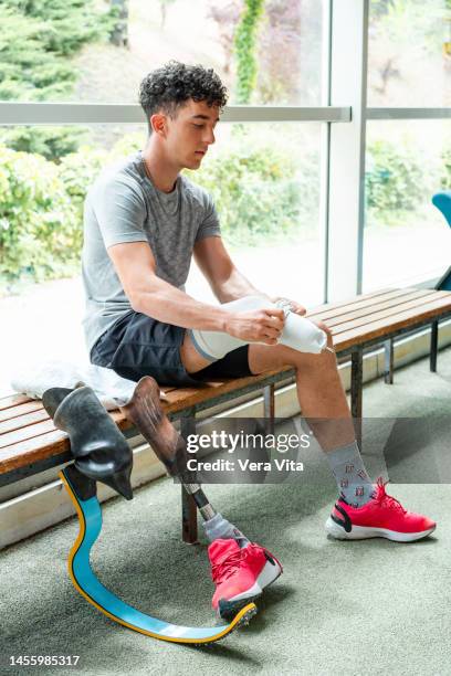 full length view of caucasian young man with lower leg amputation cleaning and changing professional prosthesitic leg at gym indoors - vita shorts fotografías e imágenes de stock