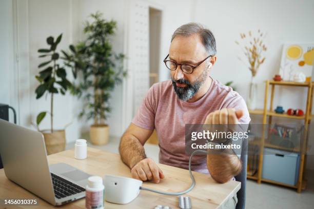 mature man checking blood pressure while having online meeting with a doctor - home medical test stock pictures, royalty-free photos & images