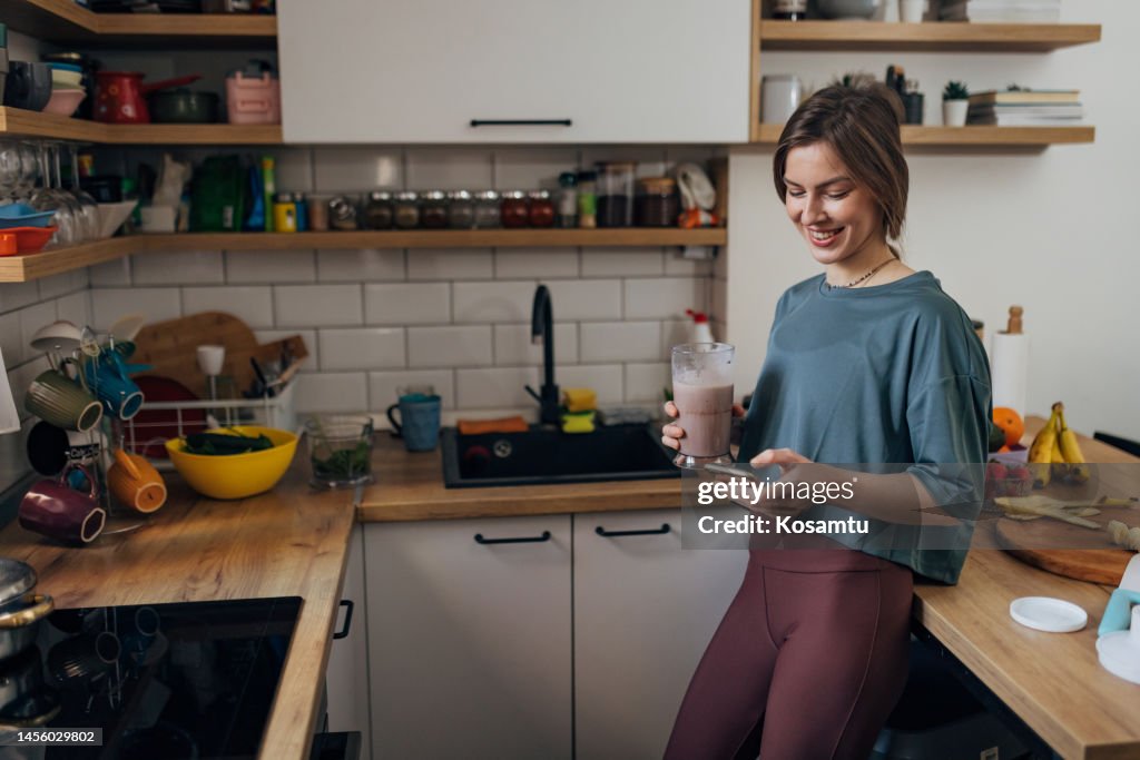 A smiling woman drinks a milkshake and uses a mobile phone while standing in the kitchen