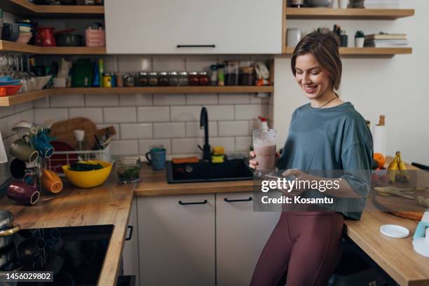a smiling woman drinks a milkshake and uses a mobile phone while standing in the kitchen - protein stock pictures, royalty-free photos & images