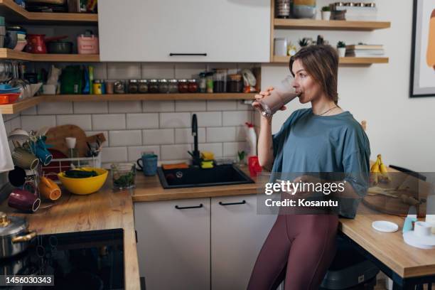 an athletic woman drinks a milkshake she made in the kitchen and surfs the internet - eiwit stockfoto's en -beelden