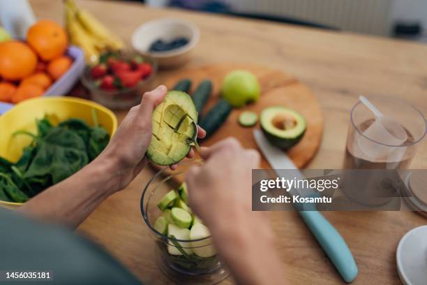 an unrecognizable woman takes the pulp from an avocado with a spoon in the kitchen - avocado smoothie stock pictures, royalty-free photos & images