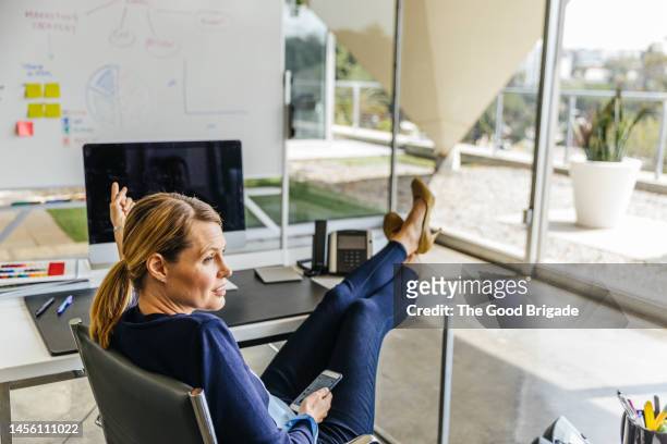 businesswoman holding smart phone while sitting with feet up on desk in office - hoher absatz stock-fotos und bilder