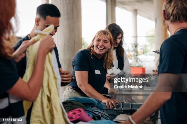 happy female volunteer looking at colleague while sorting clothes at community service center - sensibilisation communautaire photos et images de collection