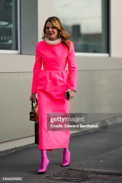 Anna Dello Russo wears golden earrings, a neon pink midi dress with corset with large ruffled collar, neon pink shiny high heeled pointed boots,...