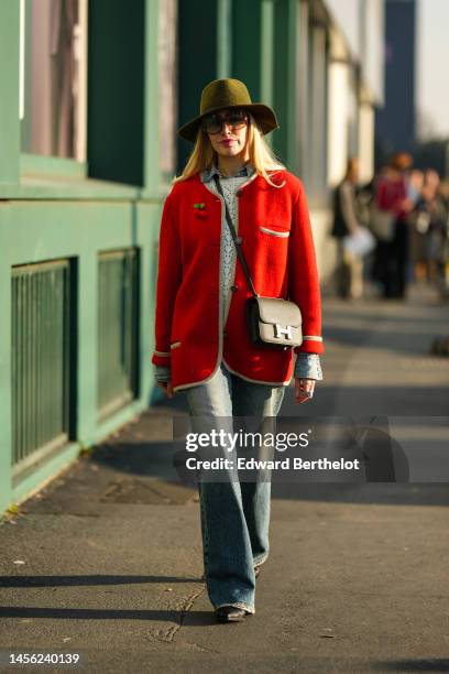 Guest wears a khaki felt / wool hat, brown sunglasses, a blue denim shirt, a white cut-out wool pullover, a red felt buttoned cardigan jacket, a...