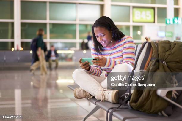 smiley african american woman at the airport using smart phone to watch a movie waiting to board the airplane. - airport phone stock pictures, royalty-free photos & images