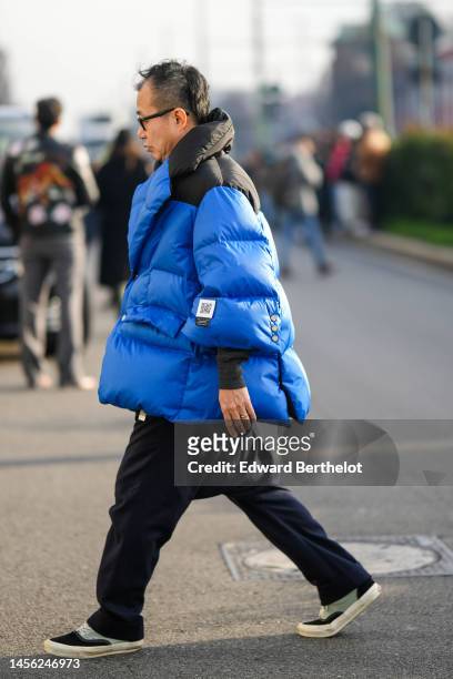 Guest wears black glasses, a black shoulder and blue oversized puffer jacket, black large suit pants, black and gray sneakers from Vans , outside...