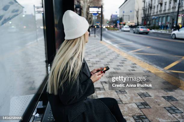 young woman with long blond hair wearing white knit hat and casual coat sitting on the bus stop and holding cell phone in hands waiting for the bus in the daytime. city life concept. - haltestelle stock-fotos und bilder