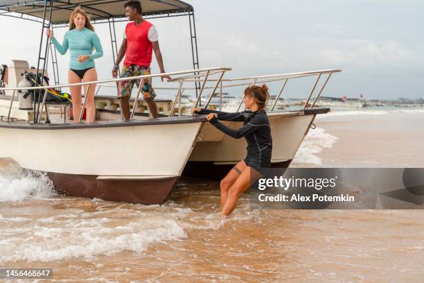 young woman holding a boat in the surf on a shorу when other members of a diving team are loading it preparing for a trip. - adventure club stockfoto's en -beelden