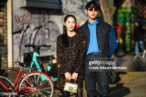 Guest wears a gold sequined long sleeves / short dress, a gold shiny leather handbag, black tights, a guest wears black circle sunglasses, a blue...