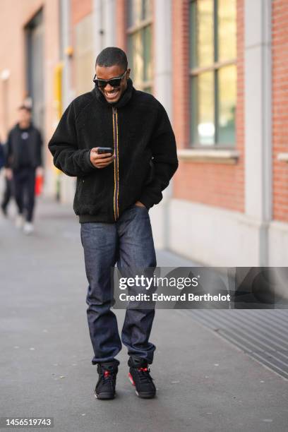 Guest wears black sunglasses, a black fluffy zipper jacket, black denim large pants, black sneakers , outside Fendi, during the Milan Menswear...