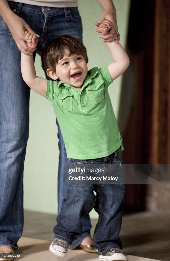 Smiling toddler boy holding his mom's hands.