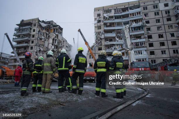 Firefighters stand near a residential building hit by a missile while their colleagues are conducting search and rescue operations on January 15,...