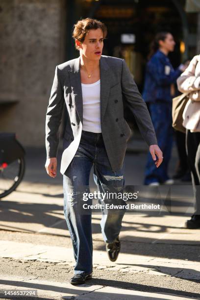 Guest wears a white tank-top, a gray blazer jacket , blue denim ripped large pants, black shiny leather derbies, a gold chain necklace , outside...