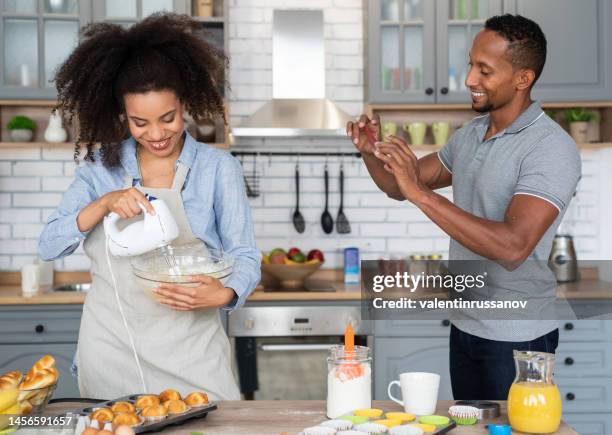 couple cooking together, having fun time in the kitchen. multiracial young woman mixing eggs in a bowl, while her boyfriend taking picture of her with his mobile phone, shooting home video or vlogging - egg icon stock pictures, royalty-free photos & images