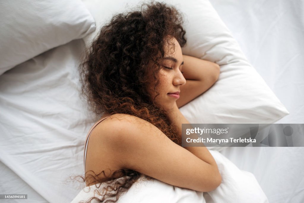 A black woman lies in bed. White linens. Dream. Cheerful morning. Good dream. Soft bed. Orthopedic mattress.