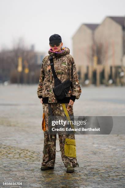 Katya Tolstova wears a black cap from Fendi, black large futurist sunglasses, silver earrings, a brown print pattern hoodie bomber coat, matching...