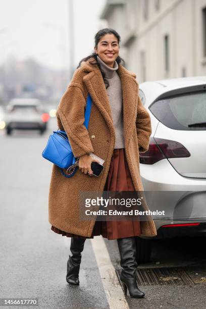 Caroline Issa wears a beige ribbed wool turtleneck pullover, a camel fluffy long coat, a brown midi skirt, a royal blue shiny leather Puzzle shoulder...