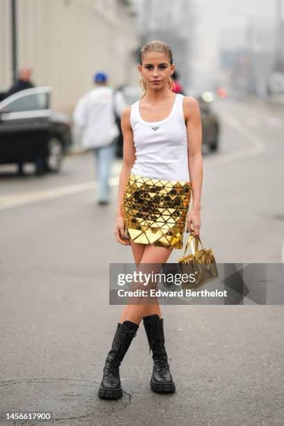 Guest wears a gold shiny varnished leather handbag from Prada, black shiny leather high laces ankle boots, a gold metallic short tube skirt , a white...