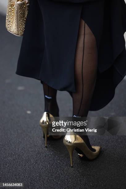 Tamu McPherson wears black dress, necklace, golden bag, heels, tights with logo print outside Prada during the Milan Menswear Fall/Winter 2023/2024...